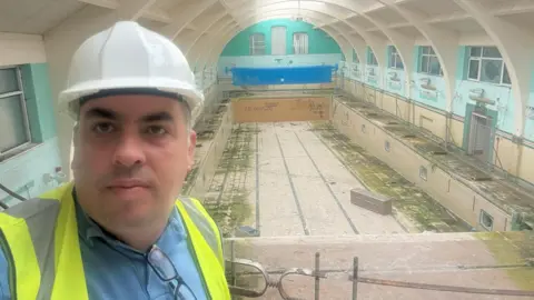 A man in a high visibility jacket is stood on a balcony overlooking an empty swimming pool in a building which has been damaged by mould, graffiti and vandalism.