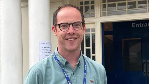 A white middle aged man with black rimmed glasses and wearing a green shirt and a blue staff lanyard smiles at the camera outside Ashcombe School. 