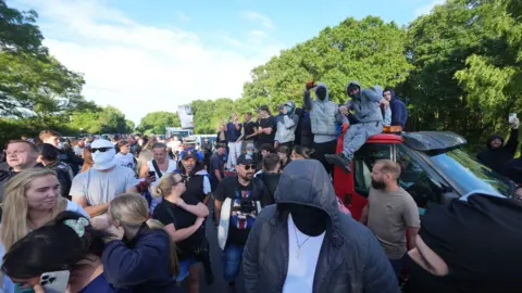 PA Media A crowd of people including some wearing full face masks or hoods - some are sitting on top of vehicles or looking at their phones. It is a bright sunny evening