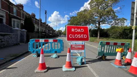 BBC Road closed sign, barriers and cones in a road, with red brick terraced homes to the left and greenery on the right. A sunny day.