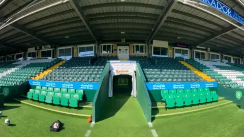 BBC The photo shows the tunnel at Huish Park and the surrounding stand in sunlight. A bag of footballs can be seen on the floor in front of the left hand dugout. 