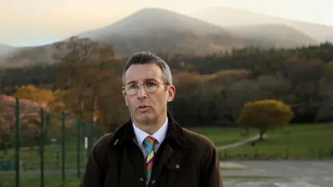 PA Media Department of Agriculture, Environment and Rural Affairs (DEARA) Minister Andrew Muir speaks about the gorse fire, at Donard Park in Newcastle, Co. Down. He has salt and pepper hair, wearing glasses, a brown jacket, white shirt and multi-coloured stripped tie. The Mournes Mountains are in the background.