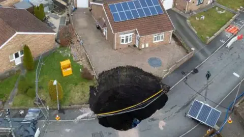 Getty Images Large sinkhole in street on the drive of one house and into the road, with other homes nearby, barriers in the street.