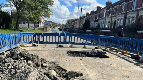A view of the hole, dug out as a square in the tarmac. A large pile of debris from it to the left. Blue barriers all around the hole in the middle of the road. Red brick terraced houses line the right of the road, trees to the left. A sunny day.
