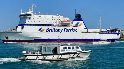 A large ferry is berthed. It is a white bird with the words Brittany Ferries  in navy blue along the side. orange lifeboats are visible on the ferry. There is a smaller white boat passing in front of it. A few passengers can be seen on the deck of the smaller vessel.