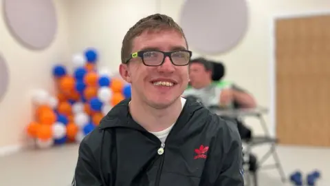 Matthew Badman looking at the camera smiling while wearing a black jacket. Behind him is another service user and lots of orange, blue and white balloons.